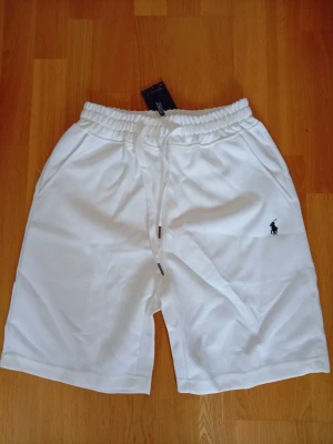 Vita shorts från Polo Ralph Lauren - Snygga vita shorts från Polo Ralph Lauren med elastisk midja och snörning. Klassisk logga broderad i marinblått på benet. Tillverkade i mjuk bomull, perfekta för sommardagar. Två sidofickor för en avslappnad look.