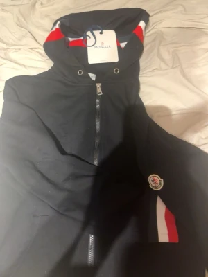 Svart Moncler hoodie med dragkedja - Snygg svart hoodie från Moncler med dragkedja framtill och huva. Detaljer i rött, vitt och blått på huvan och vid fickan samt Moncler-logga på ärmen. Perfekt för dig som gillar exklusiva streetwear-plagg med klassiska färgdetaljer.