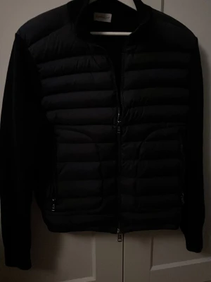 Svart cardigan moncler med stickade ärmar - Säljer en svart cardigan från Moncler med quiltad front och rygg samt stickade ärmar. Jackan har två dragkedjeförsedda fickor och en hög krage. Moncler-logga på ena ärmen ger en exklusiv touch det är inge defekter på den är i perfekt skick