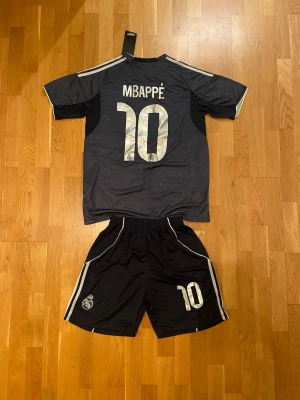 Mbappé Real Madrid bortaställ - Säljer ett svart Real Madrid matchställ med Mbappé och nummer 10 på ryggen. Tröjan har vita detaljer, klubbmärke och texten 'Emirates Fly Better' på bröstet. Shortsen har nummer 10 och klubbmärke. Perfekt för fotbollsträning eller match.
