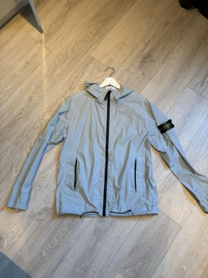 Ljusgrå vindjacka med huva Stone Island - Ljusgrå vindjacka från Stone Island med svart dragkedja, två fickor framtill och klassisk avtagbar logopatch på vänster ärm. Jackan har huva och justerbar nederkant med dragsko. Tillverkad i lätt, vindtätt material som passar perfekt för blåsiga dagar. PRISET KAN DISKUTERAS!