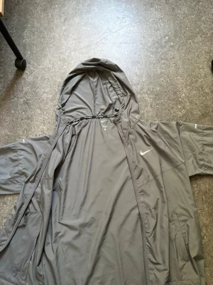 Grå vindjacka med huva från Nike - Snygg grå vindjacka från Nike med huva och dragkedja framtill. Jackan har två sidofickor och justerbar dragsko i huvan. Tillverkad i lätt och vindtätt material, perfekt för blåsiga dagar. Nike-logga på bröstet ger en sportig look.