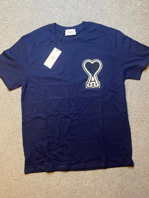 Mörkblå Ami Paris t-shirt med hjärtdetalj - Snygg mörkblå t-shirt från Ami med ett stort vitt hjärta och bokstaven A broderat på bröstet. Klassisk passform och rund halsringning. Tillverkad i mjuk bomull som känns skön mot huden. Perfekt för en clean och trendig look.