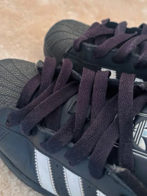 Black Adidas shoes  - Klassiska Adidas Superstar sneakers i svart läder med vita ränder på sidorna och svart tåparti i gummi. Skorna har breda svarta skosnören och den ikoniska Adidas-loggan på plösen. Perfekt för dig som gillar retrostil och streetwear.