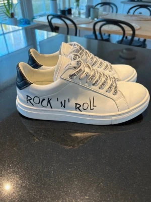 Vita sneakers fr Zadig & Voltaire i mkt fint skick, strl 37 - Snygga vita sneakers från Zadig & Voltaire  med svart häl och texten 'ROCK 'N' ROLL' på sidan. Skorna har vita sulor och vita snören med tryckt text. Klassisk låg modell med rund tå och stilren design. I mycket fint skick, endast använda ett par gånger,från djur- och rökfritt hem.