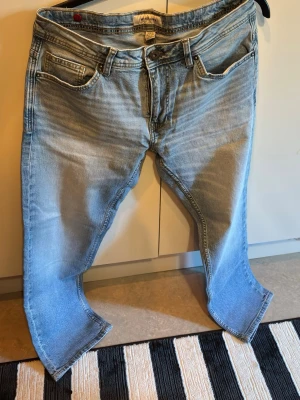 Ljusblå raka jeans - Säljer ett par ljusblå jeans med raka ben och klassisk femficksmodell. Jeansen har snygga slitningar och tvättade detaljer för en avslappnad look. Tillverkade i bomull med dragkedjegylf och knapp. Perfekta till en casual outfit.