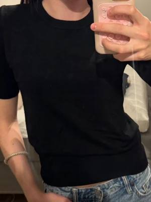Svart ribbad topp med kort ärm - En enkel och stilren svart topp med ribbade detaljer och korta ärmar. Toppen har en rund halsringning och sitter snyggt mot kroppen. Perfekt att matcha med jeans eller kjol för en clean look.
