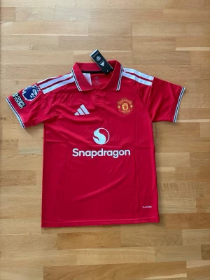 Manchester United matchtröja röd - Säljer en röd Manchester United matchtröja från Adidas med vita detaljer och klubbmärke på bröstet. Tröjan har trycket 'B. Fernandes 8' på ryggen och Premier League-märke på ärmen. Tillverkad i lätt och ventilerande material, perfekt för fotbollsträning eller match.