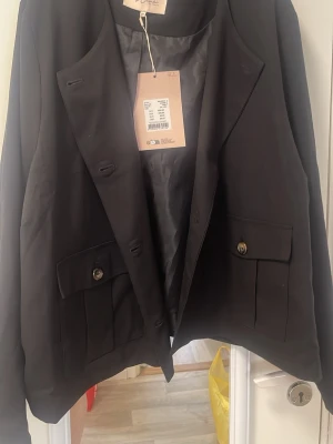 Svart klassisk kavaj med fickor - Stilren svart kavaj med klassisk passform och två stora fickor framtill med lock och knapp. Kavajen har enkelknäppning och slät insida, perfekt för en dressad look. Materialet ser ut att vara ett mjukt tyg med lätt glans.