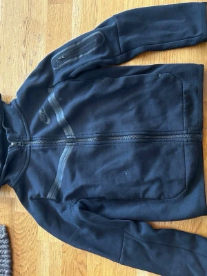 Svart Nike hoodie med dragkedja - Snygg svart hoodie från Nike med hel dragkedja framtill och diskreta svarta detaljer. Hoodien har en huva, fickor på magen och en extra ficka med dragkedja på ärmen. Tillverkad i mjukt material som passar perfekt till en sportig stil.