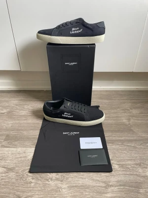 Saint Laurent skor svart - Säljer ett par svarta Saint Laurent sneakers med vit sula och handskriven logga på sidan. Skorna har klassisk låg profil, svarta skosnören och detaljer i textil. Perfekta för dig som gillar stilrena och exklusiva sneakers.