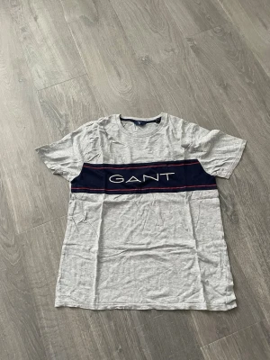 Grå GANT t-shirt med logga - Snygg grå t-shirt från GANT med korta ärmar och rund hals. Framtill finns en bred marinblå rand med vit GANT-logga och röda detaljer. Tillverkad i mjuk bomull som känns skön mot huden. Perfekt till jeans eller shorts för en avslappnad look.