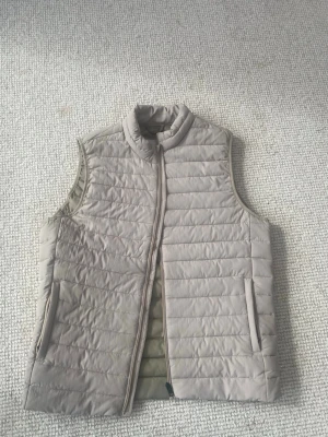 Beige dunväst från Jack & Jones - Snygg beige dunväst från Jack & Jones med quiltad design och dragkedja framtill. Västen har två fickor med dragkedja och står upp i kragen för extra stilpoäng. Perfekt att slänga över en hoodie eller tröja när det är kyligt ute.