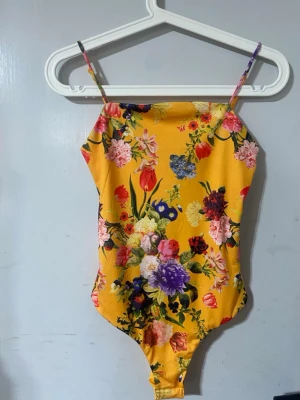 Gul blommig body från BikBok - Säljer en färgglad body från BikBok i storlek XS. Bodyn är gul med ett livligt blommönster i flera färger och har tunna axelband. Materialet känns mjukt och stretchigt, perfekt för dig som vill ha något som sticker ut.