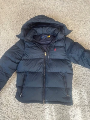 Svart dunjacka från Polo Ralph Lauren - Snygg svart dunjacka från Polo Ralph Lauren med huva och två dragkedjeförsedda fickor. Jackan har en klassisk puffer-modell och den ikoniska röda loggan broderad på bröstet. Perfekt för kalla vinterdagar och riktigt varm. Tjock jacka som inte är använd många gånger, köpt för 5999 i januari 2026