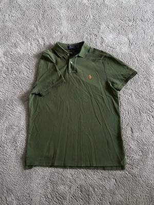 Olivgrön pikétröja från Ralph Lauren - Snygg olivgrön pikétröja från Ralph Lauren med klassisk krage och två knappar framtill. Tröjan har korta ärmar och den ikoniska broderade loggan i orange på bröstet. Perfekt till jeans eller shorts för en avslappnad look.