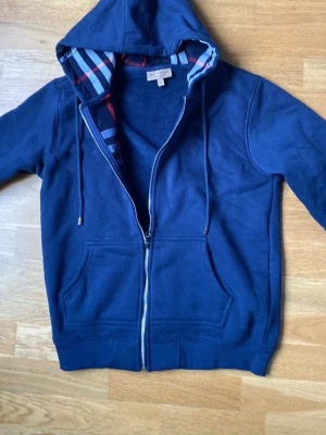 Marinblå hoodie med dragkedja - Snygg marinblå hoodie med dragkedja och huva. Insidan av huvan har ett rutigt mönster i rött, vitt och blått. Hoodien har fickor framtill och justerbara snören vid huvan. Perfekt för en avslappnad stil och passar till det mesta.
