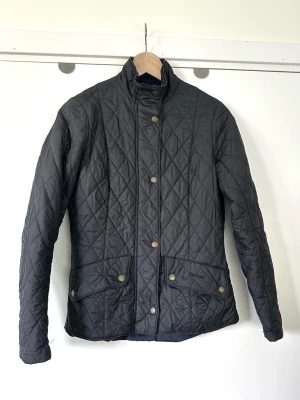 Barbour - Svart quiltad vindjacka med tryckknappar - storlek S / 36 - Stilren svart Barbour täckjacka för dam i storlek 36. Den klassiska och älskade modellen ”Flyweight Cavalry” med en smickrande passform med diamantquiltning och en ståkrage. Vackert mässingsfärgad dragkedja och tryckknappar med logotypen diskret inristat. Två framfickor med lock och tryckknappar samt en innerficka med dragkedja. Diskret broderad logotyp på ena fickan. Justerbara tryckknappar i midjan baktill för en anpassad passform. Nypris 2295:-. Mycket gott skick!