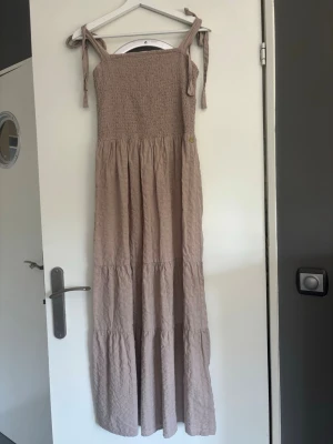 Beige långklänning med knytband - Långklänning i beige med smock upptill och axelband som knyts. Jag är 155 cm och den går ner till fötterna. Obs säljer endast via plicks köpknapp. 