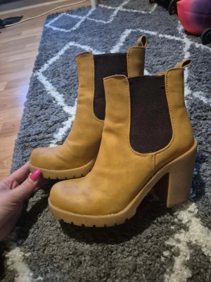 Camelbruna Chelsea boots med klack - Säljer ett par snygga camelbruna Chelsea boots med hög blockklack och grov sula. Skorna har elastiska svarta paneler på sidorna och är tillverkade i ett mockaliknande material. Perfekta för dig som vill ha både stil och höjd. Storlek 36, använda ca 5 gånger.