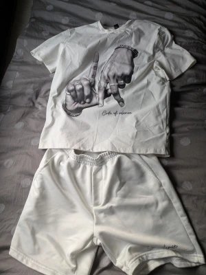 Vit t-shirt och shorts set - Säljer ett vitt set med t-shirt och shorts. T-shirten har ett tryck med två händer och texten 'Code of silence' framtill. Shortsen är enkla med resår i midjan och liten broderad text på benet. Perfekt för chill hemma eller som loungewear.