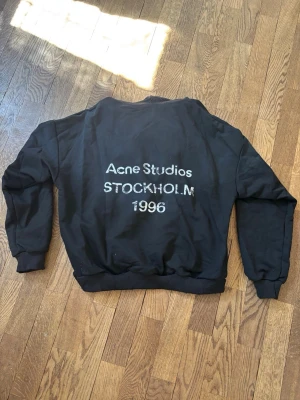 Svart hoodie Acne Studios - Svart hoodie från Acne Studios med tryck på ryggen där det står 'Acne Studios STOCKHOLM 1996' i vitt. Tröjan har rund halsringning, ribbade muddar och avslappnad passform. Perfekt för en stilren och trendig look.