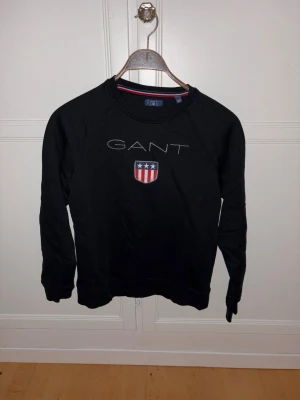 Svart sweatshirt GANT - Klassisk svart sweatshirt från GANT med rund halsringning och broderad logga samt sköld med stjärnor och ränder på bröstet. Tröjan har långa ärmar och ribbade muddar vid ärmslut och nederkant. Perfekt för en avslappnad och stilren look.