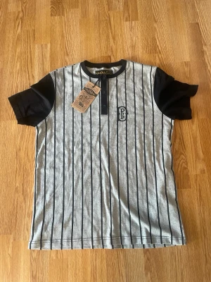 Randig t-shirt från DePalma - Snygg grå t-shirt från DePalma med svarta vertikala ränder och svarta ärmar. Modellen har knappar vid halsen och en liten logga på bröstet. Tillverkad i mjukt bomullsmaterial och har en sportig, retroinspirerad look.