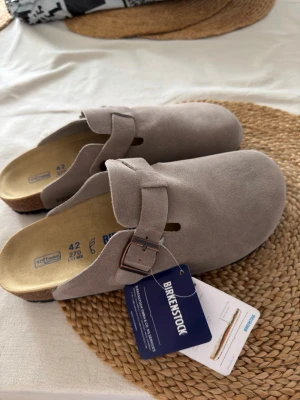 Grå mockatofflor från Birkenstock - Säljer ett par  grå tofflor från Birkenstock i mjuk mocka. Modellen har en justerbar rem med metallspänne och korksula. Säljes pga för små i storleken.