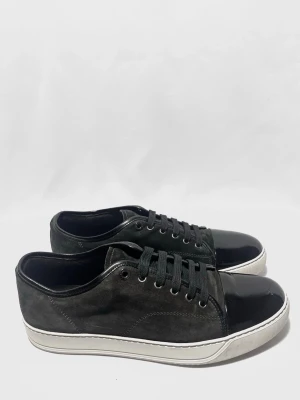 Lanvin Cap Toe Sneaker - En tidlös och eftertraktad modell från Lanvin med glansig framdel och mocka detaljer. Skorna är i mycket fint skick utan några större defekter. Färgen är mörkgrå/svart.  ✔️ Storlek: UK 9 (passar EU 43–44) ✔️ Skick: Mycket fint skick ✔️ 100 % original ✔️ Endast skorna medföljer                                                      Kontakta mig för fler frågor och funderingar!