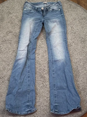 Blå bootcut jeans 14+ från Gina! - Säljer ett par ljusblå bootcut jeans med snygga slitningar och lätt tvättade partier framtill. Jeansen har klassisk femficksmodell, låg midja och vida ben. Perfekta för en avslappnad och trendig look. Använd endast ett fåtal gånger, alltså i nyskick!