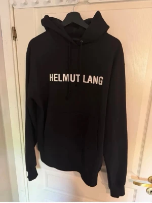 Svart Helmut Lang hoodie med tryck - Svart hoodie från Helmut Lang med vit text på bröstet. Klassisk modell med huva och dragsnören samt stor magficka framtill. Tillverkad i mjukt bomullsmaterial som känns skönt mot huden. Perfekt för en avslappnad och stilren look.