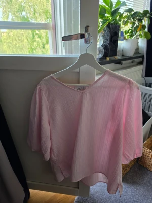 Ljusrosa blus med puffärm - Söt ljusrosa blus med v-ringning och puffiga trekvartsärmar. Tillverkad i ett tunt, plisserat tyg som ger en luftig känsla. Perfekt för dig som gillar en feminin och trendig stil.