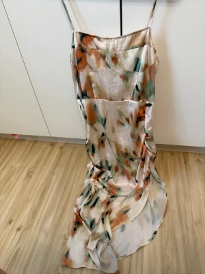 Långklänning med abstrakt mönster - Säljer en elegant långklänning med tunna axelband och abstrakt mönster i beige, grön och orange. Klänningen har en mjuk, glansig yta som påminner om satin eller silke och ett flowy fall. Perfekt för dig som vill sticka ut med en unik look.
