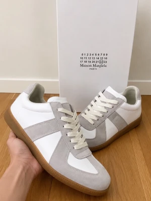 Maison Margiela vita  - Stilrena sneakers från Maison Margiela i vitt skinn och ljusgrå mocka. Klassisk låg modell med vita snören och gummisula. Perfekta för dig som gillar minimalistisk och tidlös design med exklusiv känsla.