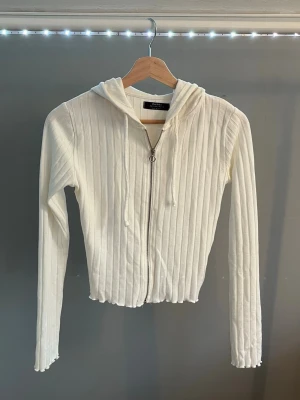 Vit ribbad hoodie med dragkedja - Säljer en vit ribbad hoodie med dragkedja framtill och huva. Tröjan har långa ärmar och är tillverkad i ett mjukt, stretchigt material. Perfekt för en avslappnad och trendig look.
