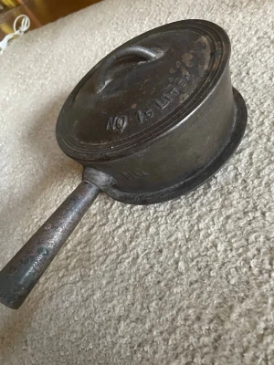 Antik gammal Gjutjärnsgryta med lock och handtag - Antik gammal Klassisk svart gjutjärnsgryta med tillhörande lock och robust handtag. Grytan har en rund form och texten 'NO 15' på locket. Perfekt för matlagning över öppen eld eller på spisen. Grytan är tung och tålig, vilket gör den idealisk för långkok och traditionell matlagning.