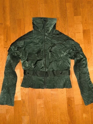Mörkgrön pufferjacka med hög krage - Snygg mörkgrön pufferjacka med hög krage och dragkedja framtill. Jackan har två bröstfickor med lock och knapp, samt quiltade detaljer på ärmar och midja. Kortare modell med markerad midja och coola sömmar för extra stil.