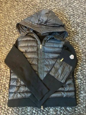Svart pufferjacka från Moncler - Snygg svart pufferjacka från Moncler med stickade ärmar och huva. Jackan har dragkedja framtill, fickor med dragkedja och Moncler-logga på ena ärmen. Materialmix av glansigt puffer-tyg och ribbstickad ull ger en modern look. Helt ny använd 2 gånger säljes då den var för liten för mig