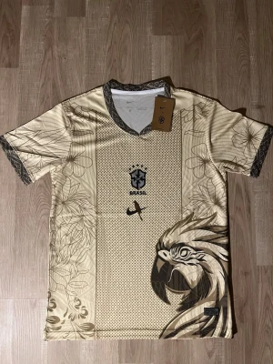 Beige Brasil Nike fotbollströja - Snygg beige fotbollströja med Brasil-logga på bröstet. Tröjan har ett unikt mönster med blommor och en stor tupp på framsidan. Svarta detaljer vid krage och ärmslut. Perfekt för dig som vill sticka ut på planen eller på läktaren. HELT NY!
