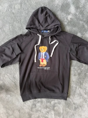 Svart hoodie Polo Bear Ralph Lauren - Svart hoodie med huva och vita dragsnören från Ralph Lauren. På bröstet finns ett tryck av den ikoniska Polo Bear i kavaj, slips och röda detaljer. Hoodien har en stor magficka och ribbade muddar vid ärmar och nederkant. Perfekt för en avslappnad och trendig stil.