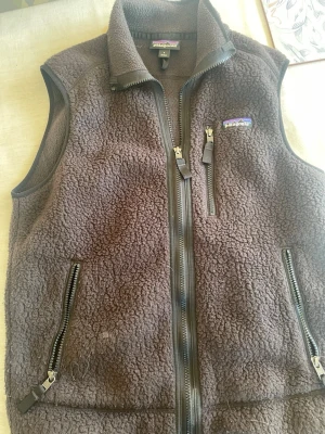 Svart väst från Patagonia - Mysig svart fleeceväst från Patagonia med hög krage och tre dragkedjeförsedda fickor. Västen har en grov, fluffig struktur och detaljer i svart. Perfekt för lager-på-lager och outdoor-vibe.