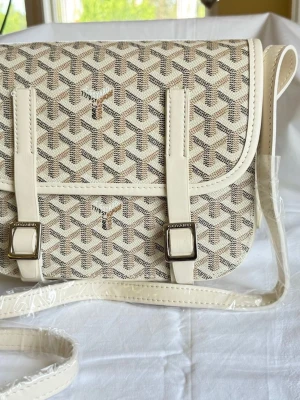 Beige axelväska med mönster - Snygg beige axelväska med klassiskt grafiskt mönster i vitt, svart och brunt. Väskan har justerbar axelrem, två dekorativa spännen framtill och detaljer i ljust konstläder. Perfekt för dig som gillar stilrena accessoarer med unik design.