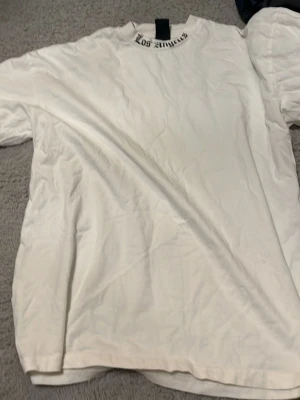 Vit t-shirt med Los Angeles-tryck - Vit t-shirt med svart Los Angeles-tryck i gotisk stil på både fram- och baksidan av halsen. Klassisk passform och rund halsringning. Perfekt för en avslappnad streetwear-look.