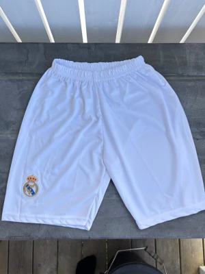 Vita Real Madrid fotbolls shorts - Storlek 176 funkar även till XS/S. Säljer ett par vita sportshorts med Real Madrid-logga på vänster ben. Shortsen har resår i midjan och är gjorda i ett lätt och snabbtorkande material, perfekta för fotboll eller träning.