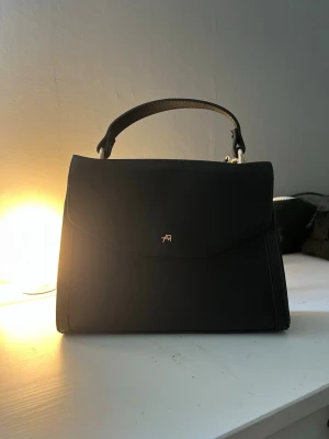 Svart stilren handväska med topphandtag - Snygg svart handväska i slätt material med ett elegant topphandtag och minimalistisk design. Väskan har ett litet guldfärgat emblem framtill och en avtagbar axelrem. Perfekt för dig som gillar klassiska och stilrena accessoarer. Välj gärna instabox🤗