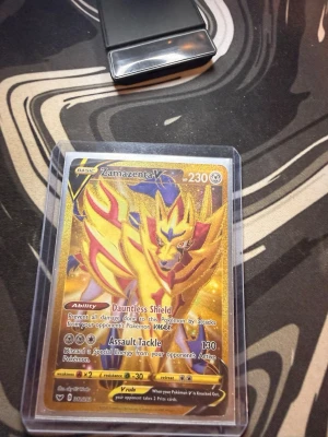 Zamazenta V Pokémonkort - Säljer ett Zamazenta V Pokémonkort med guldfärgad bakgrund och holografiska detaljer. Kortet har 230 HP och visar Zamazenta i en kraftfull pose med lila, röd och gul design. Kortet är inplastat i ett skyddsfodral.