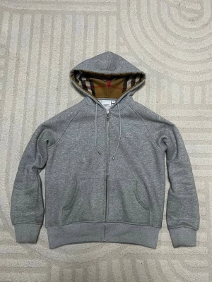 Grå zip-hoodie från Burberry - Snygg grå hoodie från Burberry med dragkedja och klassiskt rutigt foder i huvan. Mjuk bomullsblandning, två fickor fram och justerbar dragsko i huvan. Perfekt för en chill och stilren look. Den är storlek L men passar S 