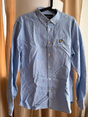 Ljusblå skjorta från lyle and Scott - Snygg ljusblå skjorta med klassisk krage och vita knappar. På bröstfickan sitter en broderad gul logga som ger en stilren touch. Skjortan har lång ärm och är tillverkad i bomull, perfekt för en clean och enkel look.