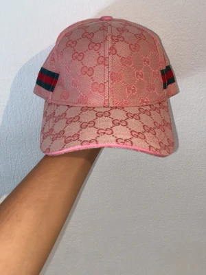 Rosa keps från Gucci med GG-mönster - Säljer en rosa keps från Gucci med klassiskt GG-monogram över hela kepsen. På sidorna finns gröna och röda ränder som ger en ikonisk touch. Kepsen har böjd skärm och justerbar rem bak. Materialet ser ut att vara canvas.